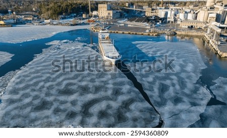 1A ice class general cargo ship manoeuvring while leaving industrial port. Sea surface covered in thin ice. Aerial front view.