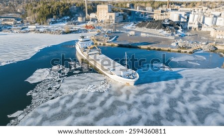 1A ice class general cargo ship manoeuvring while leaving industrial port. Sea surface covered in thin ice. Aerial front view.