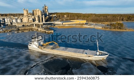 1A ice class general cargo ship manoeuvring while leaving industrial port. Sea surface covered in thin ice. Aerial side view.