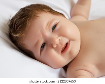 A 1-month-old Baby Is Lying And Crying Because Of Feeling Hungry On A White Mattress. The Infant Feels Unwell Due To Sleepiness. Newborn On White Blanket. Beautiful Portrait Of Little Child Girl