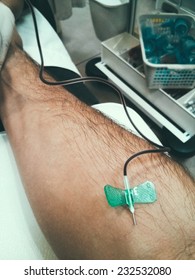 1st Person POV View Of A Right Arm With A Needle Stuck In It; Taking Blood For A Blood Test. In The Background Medical Equipment Can Be Seen, Indicating It Is In The Nurse's Room.