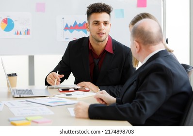 2 Adults caucasian businessmen are talking. A focused businessman is holding pen and look at a teammate and have brown cup, book, laptop put on desk and whiteboard are back with more graph on paper. - Shutterstock ID 2174898703