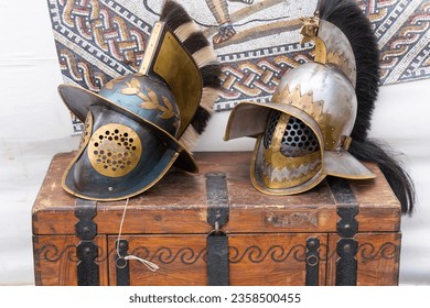 2 ancient roman gladiator helmets on a brown wooden chest with a mosaic image in the background