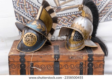 2 ancient roman gladiator helmets on a brown wooden chest with a mosaic image in the background