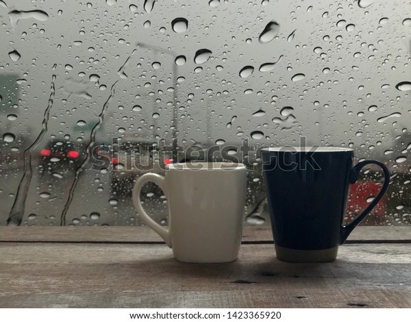 2 Coffee mugs on old wooden planks,with dimly\
light, in front of window with raindrops,blurred light,traffic on\
the road in the city,colorful bokeh as outside view.Coffee time in\
rainy day concept.