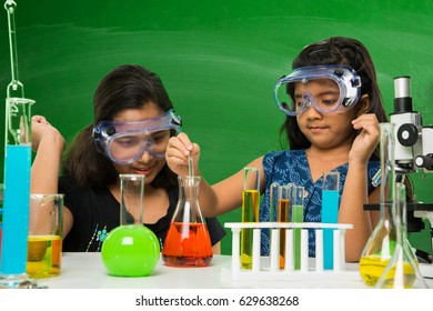 2 Cute Little Indian Girls Doing Science Experiment Or Project In A Classroom With Green Chalkboard Having Science Doodles