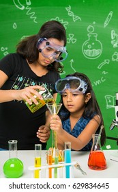 2 Cute Little Indian Girls Doing Science Experiment Or Project In A Classroom With Green Chalkboard Having Science Doodles