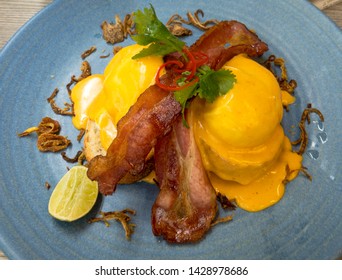 2 Eggs Benedict With Crispy Bacon And Fried Onions On A Blue Plate