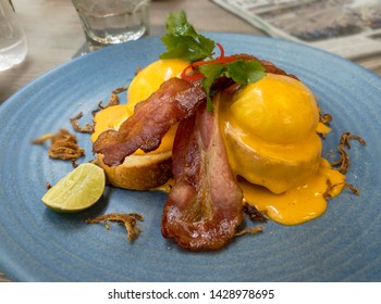 2 Eggs Benedict With Crispy Bacon And Fried Onions On A Blue Plate