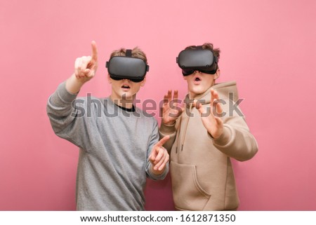 2 emotional men in VR helmets isolated on pink background. Two funny guys are wearing VR helmets on a pink background, looking surprised at the camera and pointing their finger.