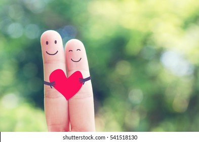 2 Fingers Holding Red Heart While Smiling Face With Nature Bokeh Background. Copy Space. Happy Couple In Love.