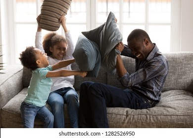 2 Happy Little Children Laughing Enjoying Funny Pillow Fight With African American Dad Sitting On Sofa, Happy Joyful Kids Daughter Son Playing With Father, Black Family Having Fun With Daddy At Home