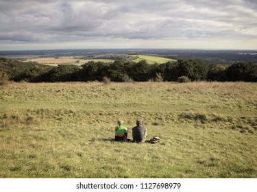 2 people sitting on the grass looking out onto a green English countryside landscape with some moody clouds forming in the sky - Shutterstock ID 1127698979