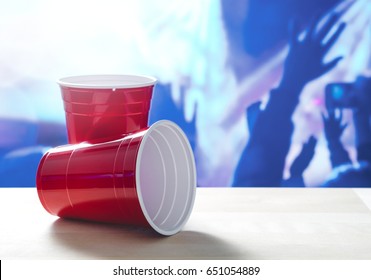 2 Plastic Red Party Cups On A Table. One On Its Side. Nightclub Or Disco Full Of People Dancing On The Dance Floor In The Background. Perfect For Marketing And Promotion For Events Or College Fest.