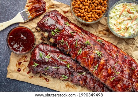 2 racks of ribs sitting on brown paper with barbeque sauce brush, sides of cole slaw, baked beans and garnished with fresh thyme herbs