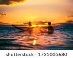 2 silhouette surfers sitting on surfboards in the ocean during a perfect Japanese sunrise. The suns rays are like a star & the colours are very vivid.