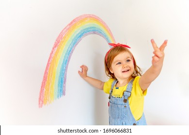 2 years old girl is having fun at home. Sign of hope. Coronavirus. Stay at home. Indoor kids activities. Girl in colorful clothes is drawing rainbow on the wall. Baby girl is showing finger.