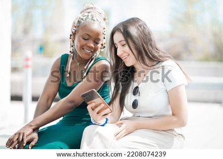 2 young multiethnic girls sitting on a bench outside and watching funny content on the smartphone