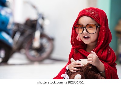 20 months old cute Indian baby girl wrapped in dark red scarf sitting and smiling innocently while holding a money toy in hands