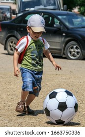 2011.05.29, Maloyaroslavets, Russia. A Little Cute Boy In A Baseball Cap And Backpack Runs In The Yard Playing A Big Soccer Ball.