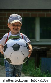 2011.05.29, Maloyaroslavets, Russia. A Little Cute Boy In A Baseball Cap And Backpack Runs In The Yard Playing A Big Soccer Ball.