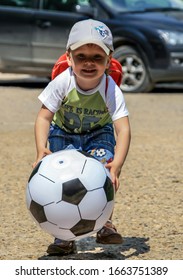 2011.05.29, Maloyaroslavets, Russia. A Little Cute Boy In A Baseball Cap And Backpack Runs In The Yard Playing A Big Soccer Ball.