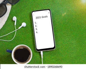 2021 Goals Checklist. Smartphone With 2021 Goals List, Earphones, Coffee, Hat And Sunglasses On Green Grass Background.