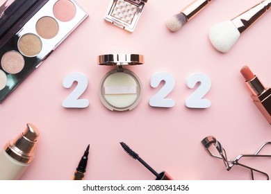 2022 Beauty cosmetic makeup products trends concept. Top view of 2022 white number with powder, lipstick, foundation, eyeshadow, brush, eyeliner and mascara on pink background.