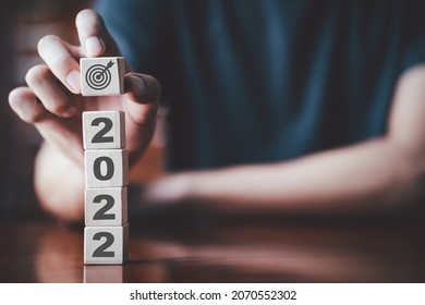 2022 New Year Setting Goals And Achievements This Year, Starting And Business, Financial Planning Work Concept. Man Hand Holding Wooden Cube Marked With A Target Board On Stack Of 2022 Cubes.