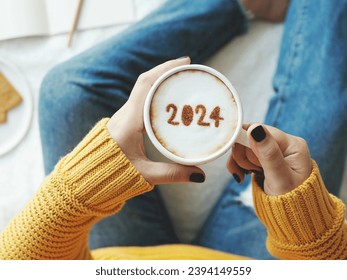 2024 new year's goal setting, number 2024 on frothy surface of cappuccino in white coffee cup holding by woman in yellow knitted sweater with jeans sitting on bed with white blanket background.