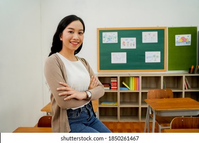 20s Asian female young adult teacher with arms folded in elementary school building in background. Happy smiling confident educator in classroom with desks and blackboard. Occupation concept