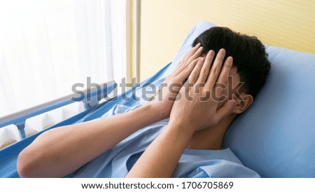 20s Asian patient young male anxious, stress and sensitive feeling from quarantine so raise hand palm to cover face on his bed in sickroom after admitted at hospital for long days