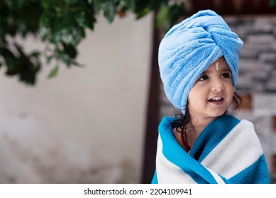 22 Months Old Cute  Indian Baby Girl Wearing Blue Hair Towel Wrap Turban And Body Wrapped With Blue And White Towel, Sandalwood Anoit On Her Forehead Sitting And Smiling.