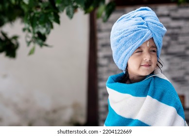 22 Months Old Cute  Indian Baby Girl Wearing Blue Hair Towel Wrap Turban And Body Wrapped With Blue And White Towel, Sandalwood Anoit On Her Forehead Sitting And Giving Innocent Expressions.