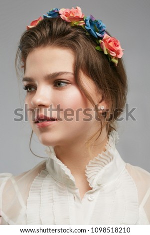 2/3 view portrait of a pretty young lady with a boho flower crown, in a white vintage blouse. The romantic girl looking pensively aside on the grey background, peach pink and blue roses in her hair.