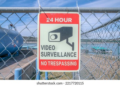 24 Hour Video Surveillance And No Trespassing Signage On A Chain Link Fence- Lake Austin, Austin, TX. Close-up Of A Signage Against The View Of Tanks And Lake At The Back.