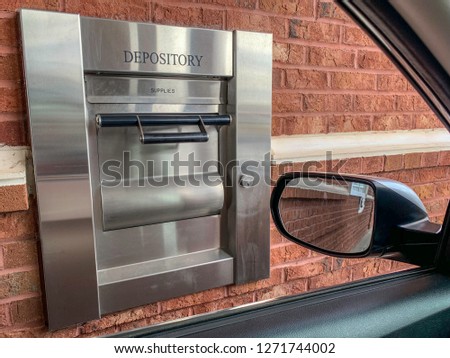 The 24-hour drive-thru night depository terminal of a neighborhood bank set on red brick wall and framed through portion of a car.