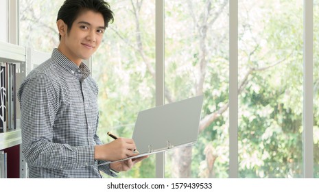 A 25-35 Year Old Asian Business Man Smiled Happily In The White Office. He Was Checking The Documents To Sign The Trade Documents.