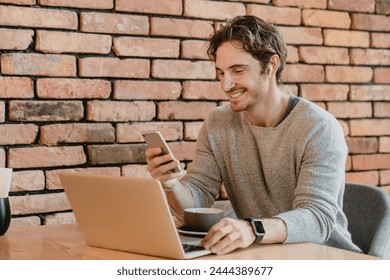 25s smiling attractive businessman working on his laptop and phone in loft cafeteria. Caucasian freelancer multitasking, performing online meeting, doing homework project report - Powered by Shutterstock