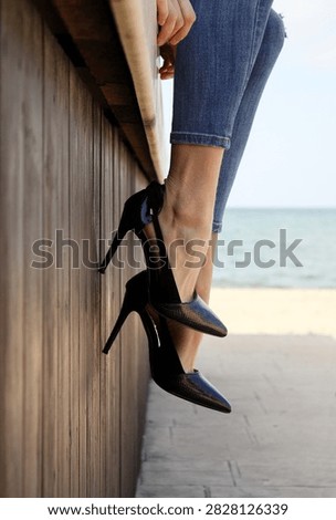 29 July at exactly 12:48 PM. A uniquely captivating cinematic frame from a low angle, documenting an elegant moment brimming with urban poise and coastal grace. A woman's legs, clad in fitted denim jeans with a textured, rugged weave, dangle gracefully while wearing luxurious, sleek high-heeled stilettos in a deep black shade with a subtle snakeskin pattern that catches the light with a mesmerizing sheen. She is perched on a raised ledge, her legs dangling elegantly beside a vertical wooden panel wall whose grain and prominent lines emphasize the warm, tactile details of natural timber, while the background stretches out into a bright, crisp seaside horizon blending seamlessly with a serene sandy beach, crafting a visual masterpiece that defies imagination.