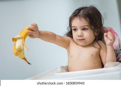 2-year-old Latina baby girl sitting in a highchair, holding a banana in her open hand and bitten, is eating.