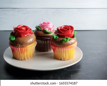 3 Cupcakes Sitting On A Plate, Yellow And Chocolate With Chocolate Frosting And Red And Pink Roses Made From Frosting, On A Black Slate Surface With A Gray Shiplap Background, Festive And Delicious.