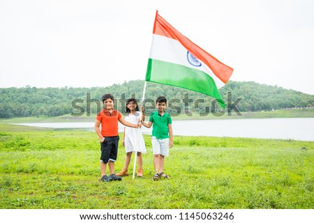 3 cute little indian kids holding, waving or running with Tricolour near a lake with greenery in the background, celebrating Independence or Republic day  