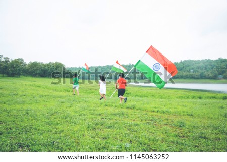 3 cute little indian kids holding, waving or running with Tricolour near a lake with greenery in the background, celebrating Independence or Republic day  