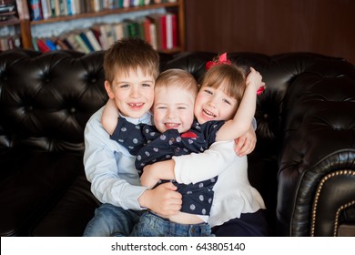 3 Happy Children, Two Brothers And A Sister (2 Boys And A Girl) 3-7 Years Old Are Sitting On A Black Leather Sofa Hugging, Hugs, Gay Smiles