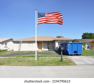 30 Yard Blue Dumpster And American Flag In Front Of Suburban Fire Damaged Ranch Style Home All Boarded Up On Clear Blue Sky Day In USA