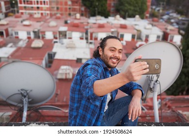 30 Year Old Man Taking A Picture With His Cell Phone And In The Background Urban Landscape Of Buildings Taken From A High Point Of View.