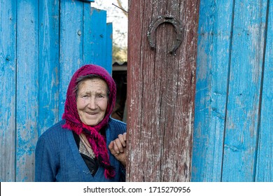 30.04.2018 Sutiski Ukraine An old woman in a scarf. Portrait of a lonely old woman standing in a village next to the blue fence on which an old horseshoe is nailed. Entrance.
