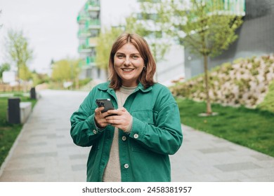 30s cheerful woman tourist dressed in casual look holding smartphone gadget in hand for communicate on the city street. Happy woman chatting on phone and relaxing outdoors. Urban, people concept. - Powered by Shutterstock