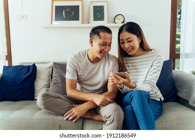 30s Young Adult Asian Man And Woman Playing A Mobile Phone Together Sits On Sofa In Cozy Living Room At Home. Happy Couple Internet User In Casual Clothes On Couch. Technology Usage In Asia Concept